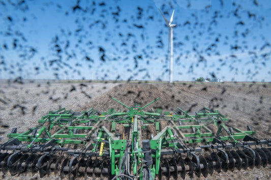 Spring fieldwork from the tractor seat, looking through a dusty cab window at farm equipment, open Iowa fields, and wind turbines in the distance.