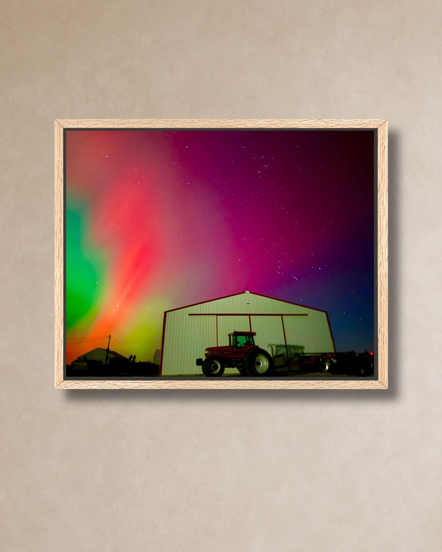 Natural oak framed canvas print of northern lights over a rural farm shed and tractor, shown on a neutral wall.