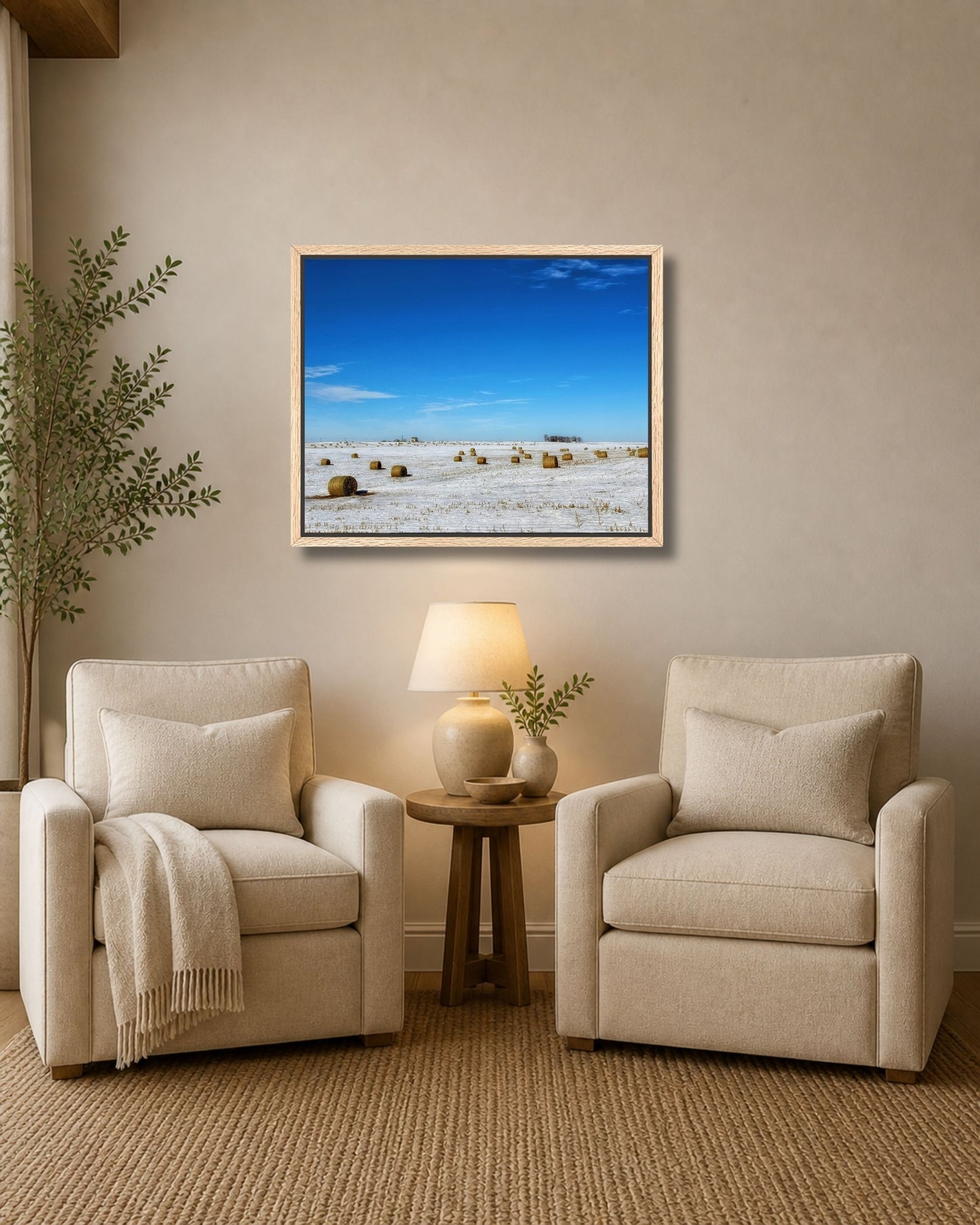Natural oak framed winter farm landscape wall art displayed between two cream armchairs in a soft neutral sitting area.