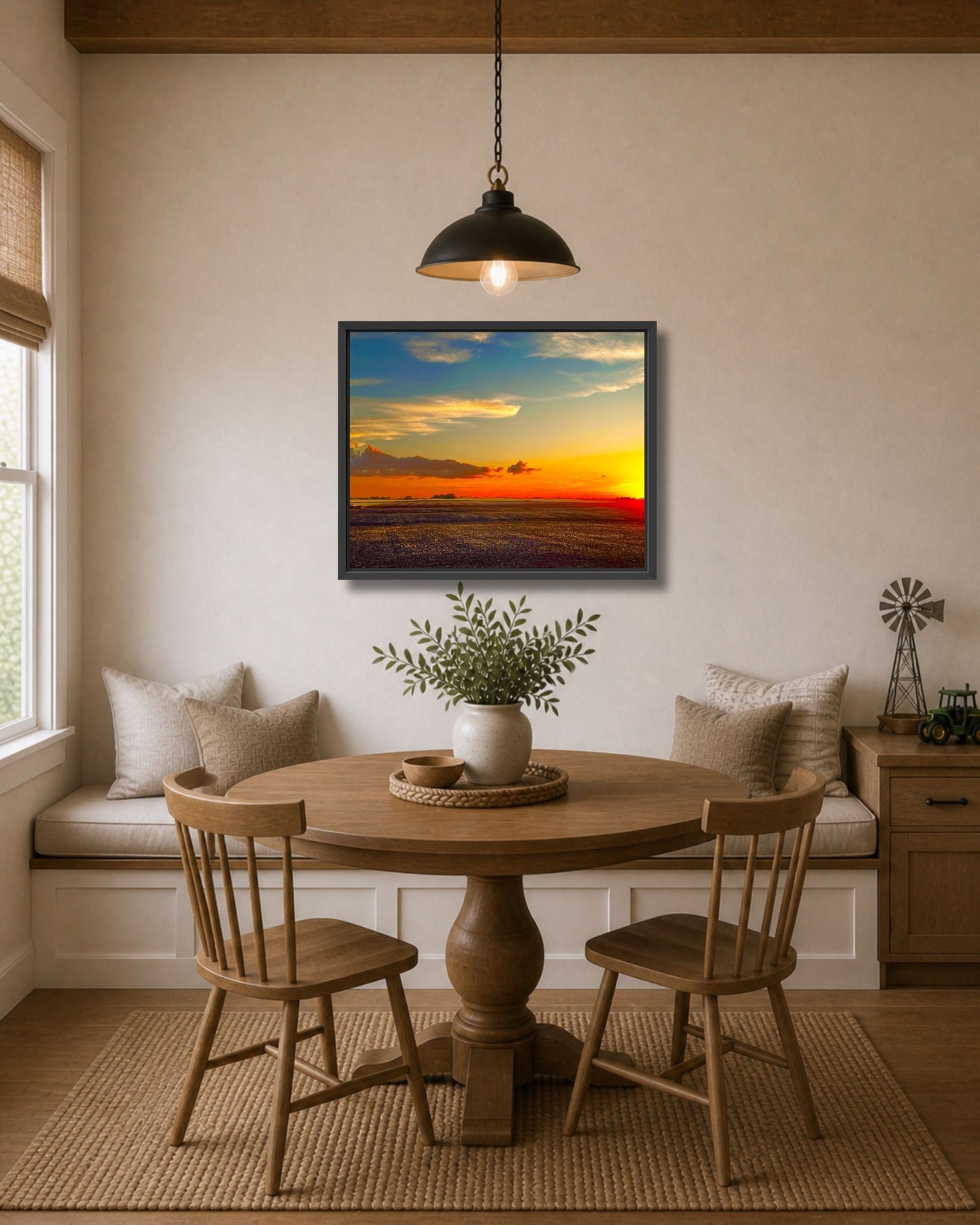Black framed farmland sunset wall art displayed above a round wood dining table in a cozy farmhouse nook.