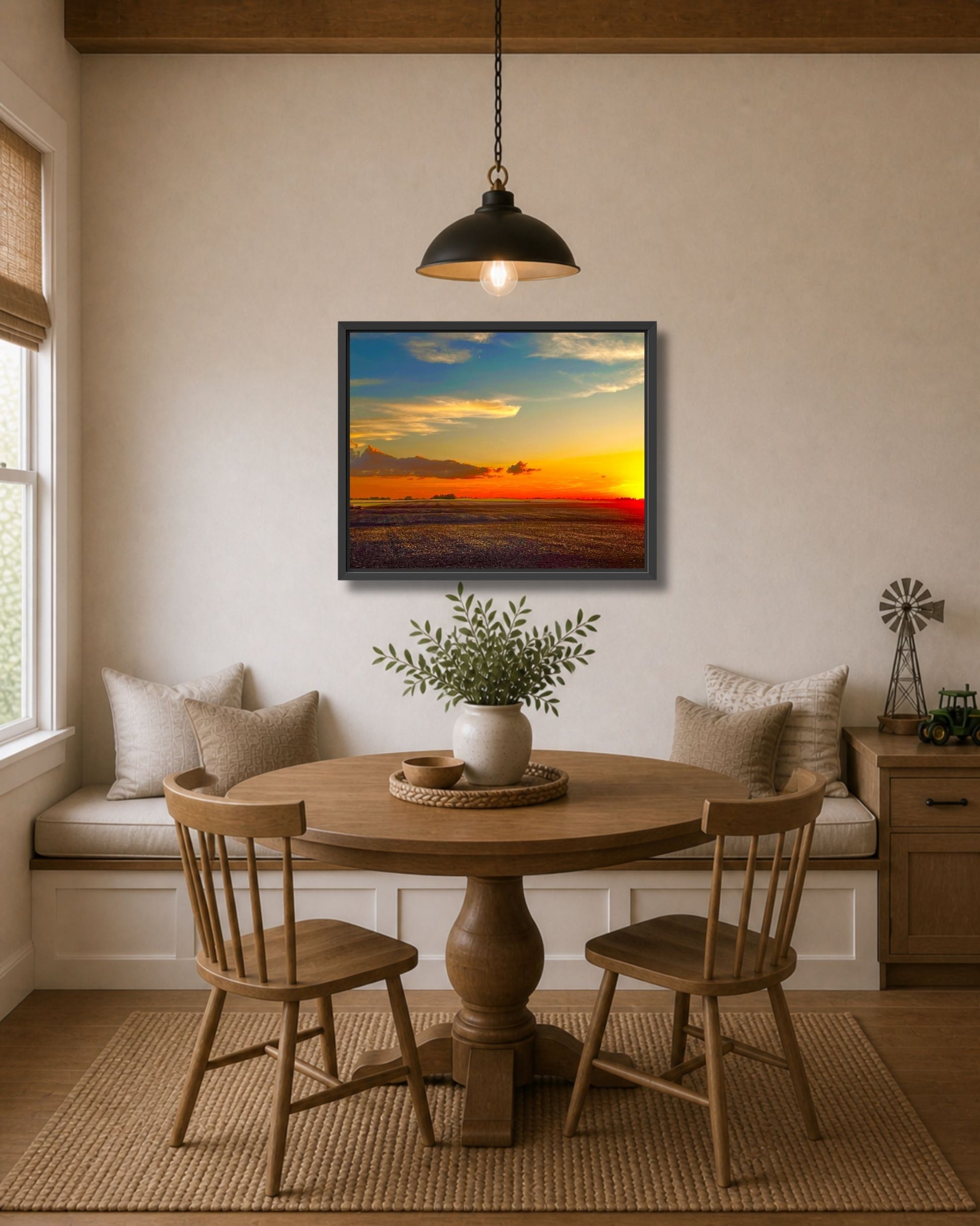 Black framed farmland sunset wall art displayed above a round wood dining table in a cozy farmhouse nook.